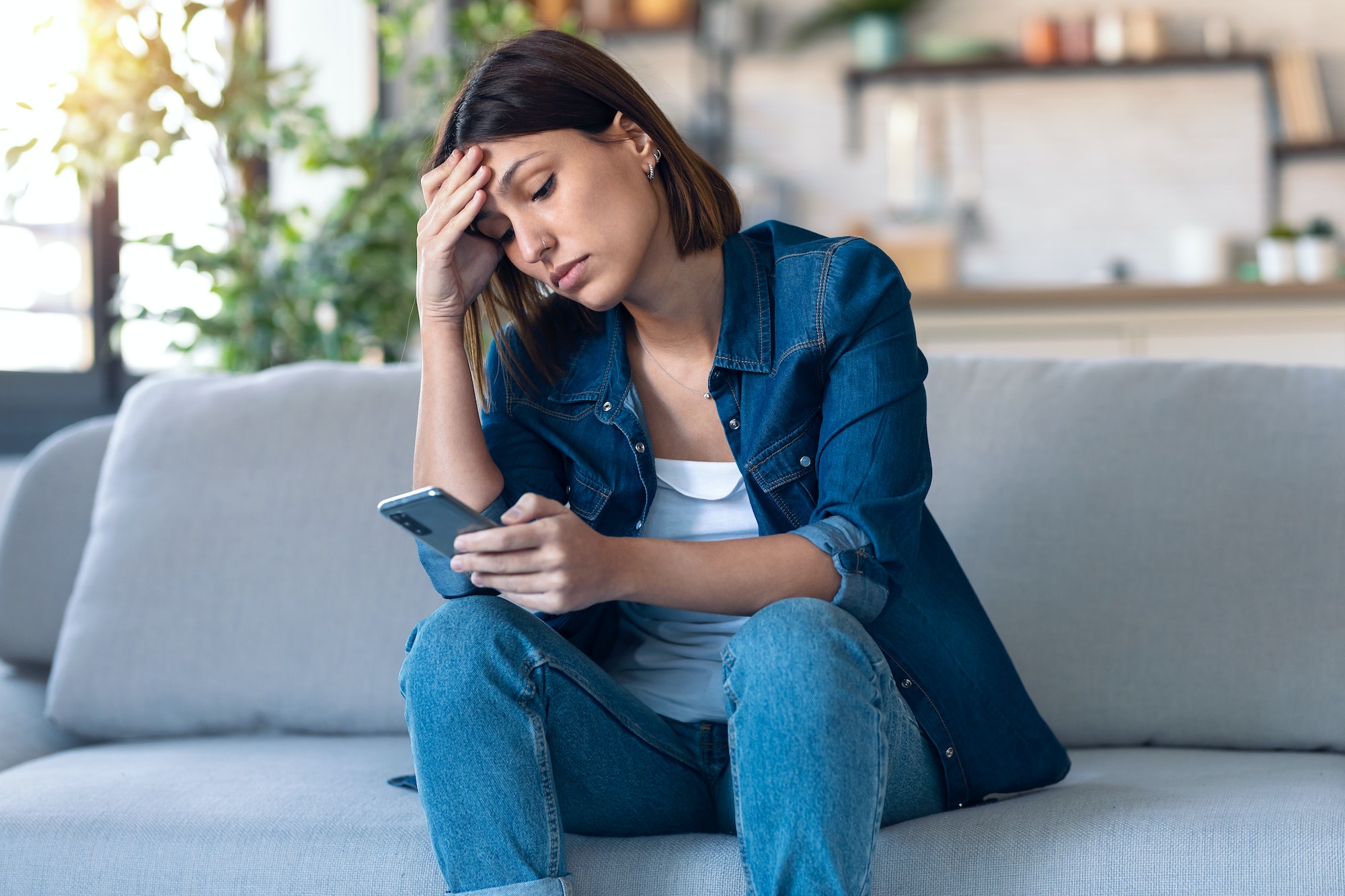 Worried young woman using her mobile phone while thinking about problems sitting on couch